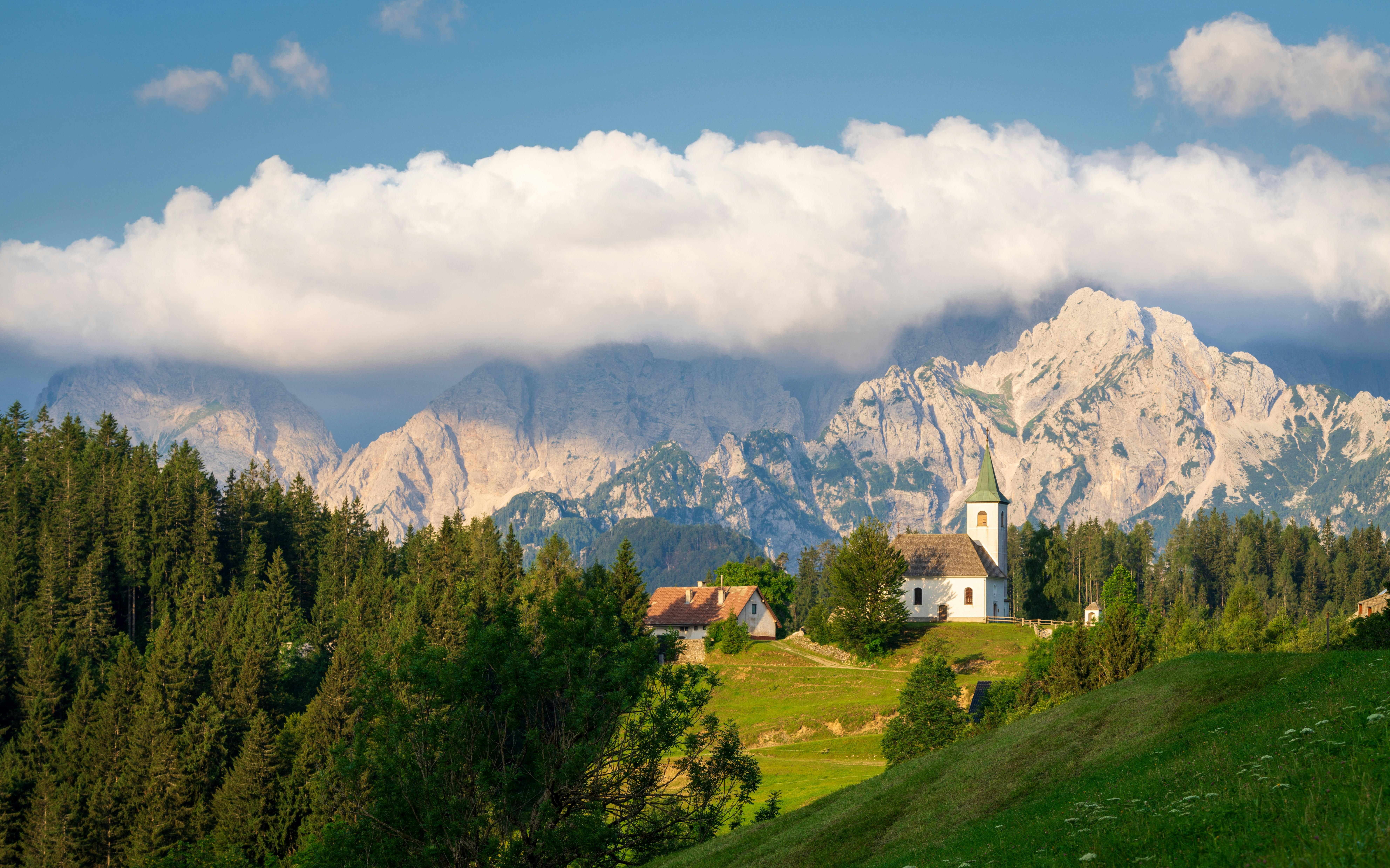 A white church and farmhouse on a green hillside below the Alps along the Solčava Panoramic Road